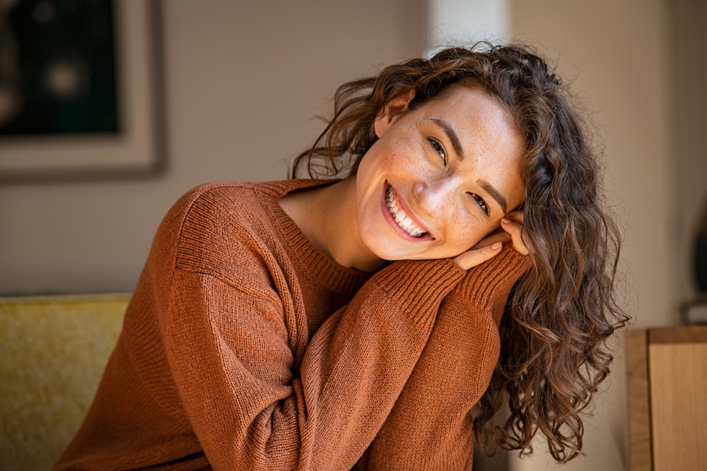 A young professional woman is smiling, showcasing her clear Invisalign aligners, almost invisible, while working on a laptop in a sunlit coffee shop near Needham, MA. No text on the image.